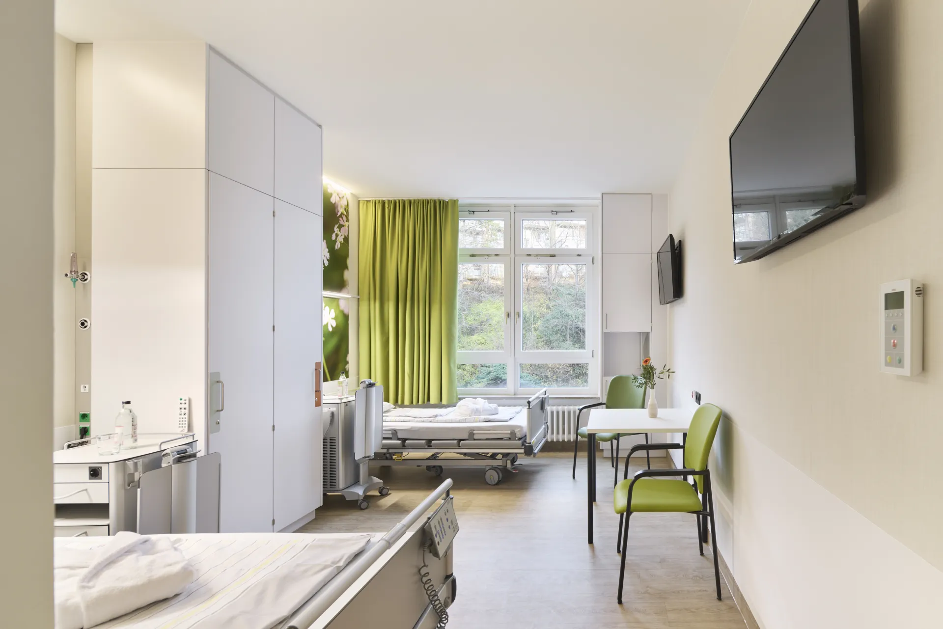 Helles Patientenzimmer mit zwei Betten, grünem Vorhang, zwei grünen Stühlen, zwei Wandfernsehern und großem Fenster mit Blick ins Grüne